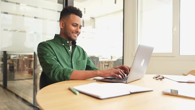 Successful freelancer. Happy millennial Indian man sitting at desk with laptop, smiling and typing, enjoying remote work, male student studying on the distance