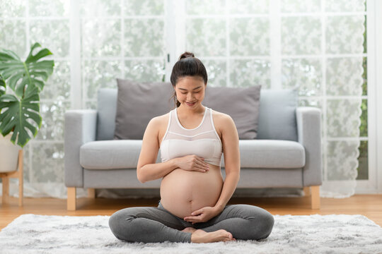 Happy Pregnant Asian Woman sitting on bed holding and stroking her big belly at home,Pregnancy of young woman enjoying with future life relax at home,Motherhood and Pregnant Concept,Soft focus