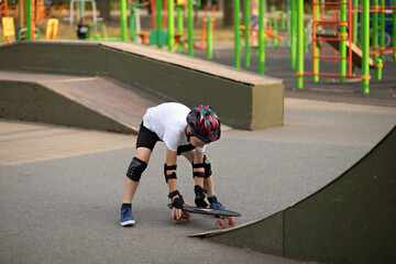 Cute kid boy child in a helmet sitting in a special area in skatepark and standing on skateboard. Summer sport activity concept. Happy childhood.
