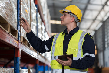 Male warehouse worker wearing uniform checks stock inventory in warehouse. Male worker holding clipboard and checking barcodes on boxes on shelf pallet in storage warehouse - obrazy, fototapety, plakaty