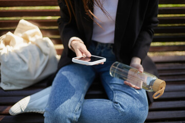 Young female sitting on a bench with a smart phone and glass water bottle in hands. Unrecognizable girl in blue jeans chilling outdoors and drinking fresh water from a reusable glass bottle