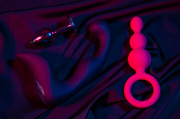 Black prostate stimulator, metal butt plug and pink anal beads in pink neon on a black silk sheet. - obrazy, fototapety, plakaty