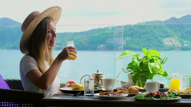 Smiling woman at cafe table enjoys glass of orange juice with sea view. Breakfast in hotel on summer holidays in tropical resort. Phuket island, Thailand, Asia.