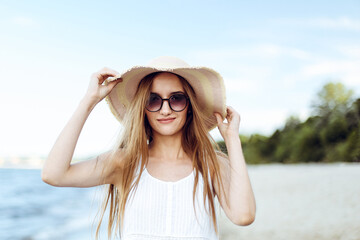 Happy smiling woman in free happiness bliss on ocean beach standing and posing with hat and sunglasses. Portrait of a female model in white summer dress enjoying nature during travel holidays vacation