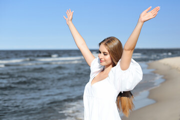 Happy, beautiful woman on the ocean beach standing in a white summer dress, raising hands.
