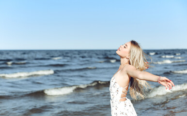 Fototapeta na wymiar Happy blonde beautiful woman on the ocean beach standing in a white summer dress, open arms.
