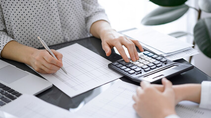 Woman accountant using a calculator and laptop computer while counting taxes with a client or a colleague. Business audit team, finance advisor.