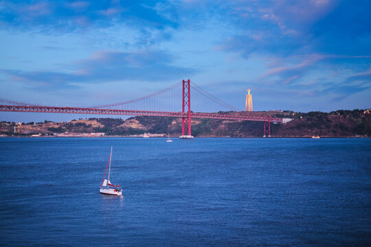 View of 25 de Abril Bridge famous tourist landmark over Tagus river, Christ the King monument and a yacht boat in the evening twilight. Lisbon, Portugal