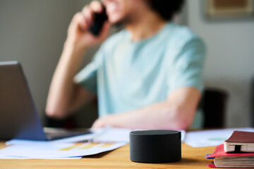Close-up of smart speaker standing on table with young man talking on mobile phone in background