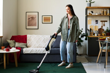 Young woman in headphones listening to music and doing housework with vacuum cleaner in the room - obrazy, fototapety, plakaty
