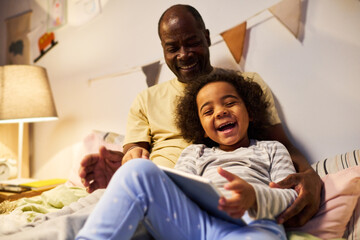 Happy dad watching funny video on digital tablet together with his little son before sleep in...