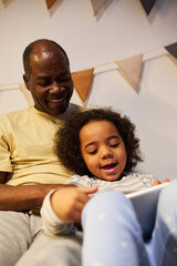 Vertical image of African American dad using tablet pc together with his son in bed