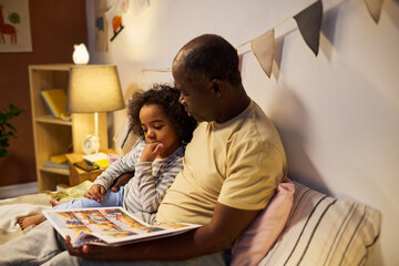 African American dad reading fairy tale to his child, they sitting on bed and watching pictures in...