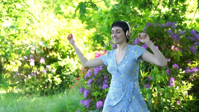 Outdoor portrait of happy  woman in blue dress and headphones dancing in the public park. Listen to online music anywhere and be yourself concept