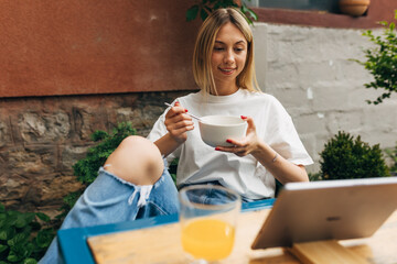 Front view of a Caucasian woman having a breakfast outside while watching a TV show on tablet.