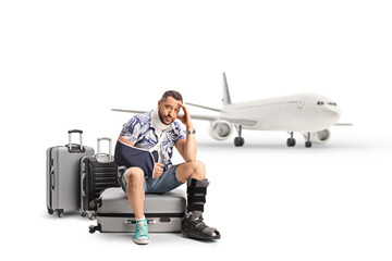 Sad male tourist with an injured arm and leg sitting on suitcases in front of an airplane