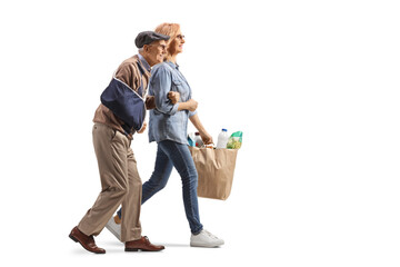 Full length profile shot of a woman helping an elderly man with an injured arm and carrying grocery...