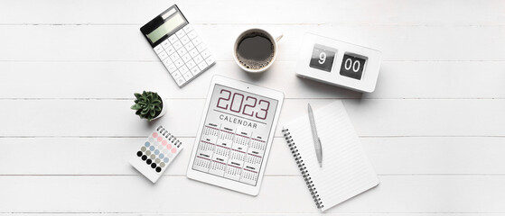 Calendar for 2023 year on screen of tablet computer, notebooks, alarm clock, coffee and calculator on white wooden background