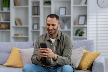 Fototapeta premium Young smiling African American man sitting on sofa at home and using phone, typing messages, texting, playing online games