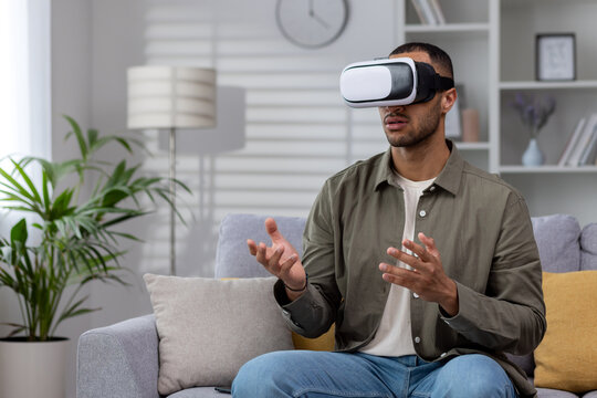 African american young man sitting on sofa at home wearing 3d glasses, wearing virtual reality glasses, playing online games