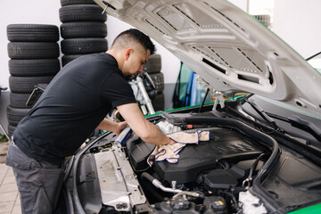 Side view of a male mechanic working in his garage. Young bearded man checks the engine in the car, the hood is open