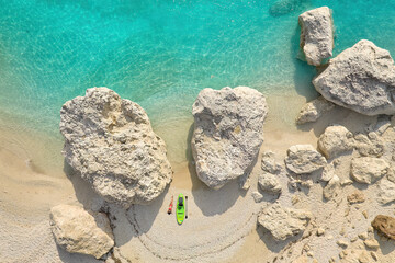 Fototapeta na wymiar A perpendicular aerial view of a hidden sandy beach and a young woman in a red swimsuit lying next to a green paddleboard. Green-blue clear sea, sunny day, big rocks, Lefkada, Greece. 