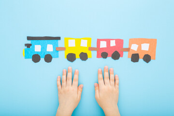 Little child hands and colorful paper train with wagons on light blue table background. Pastel color. Point of view shot. Closeup. Top down view.