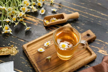 Board with cup of hot chamomile tea and scoops on black wooden background