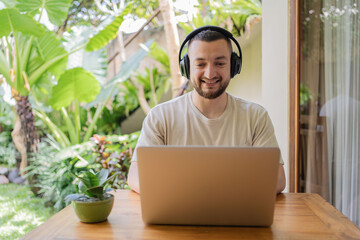 Young man with beard in casual clothing working on laptop with headphones while sitting at table in...