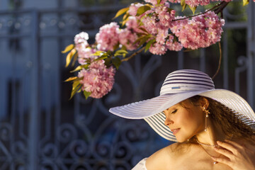 A beautiful girl in a white short airy dress and a white wide-brimmed hat on vacation near cherry blossoms