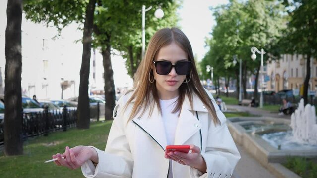 a young beautiful girl is standing on the street smoking and typing a message on her red phone 4k 
