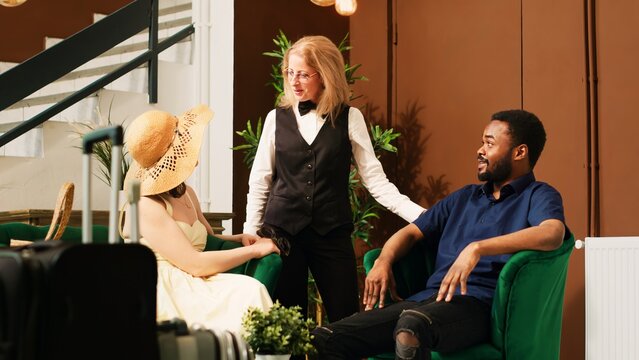 Resort employee chatting with people in lobby, couple waiting to do registration process in lounge area. Hotel guests asking worker about leisure activities, summer vacation. Handheld shot.