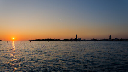 Sunset over the Venetian lagoon with silhouettes of historic buildings and calm water reflecting the warm evening light, Venice, Italy © Filippo
