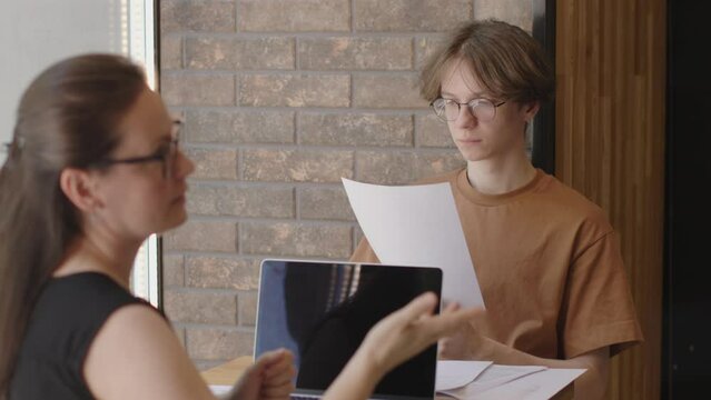 young male student guy in brown t-shirt sits carefully reads paper documents while female project manager HR discusses problems and gesticulates, job interview, hiring employee office work concept