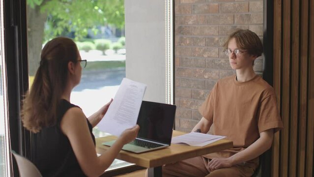woman in black interview young male student in office. first job application female manager sits table laptop looks through paper documents, man opposite nervous attentively waiting decision.