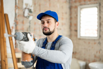 Portrait of positive man builder posing with handheld demolition hammer at indoor construction site