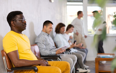 Middle-aged male patient sitting on the bench in waiting room of clinic together with other guys