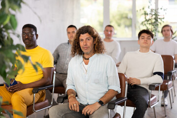 Positive stylish adult man hipster with long wavy hair listening with attention to lecture, sitting...