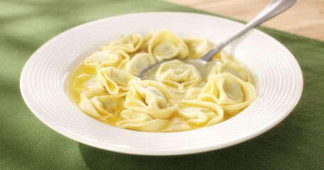 tortelloni with ricotta and spinach in chicken broth with spoon. hot italian pasta on a white plate. ready to eat food. green background. mediterranean cuisine. sunny day. angle view. 