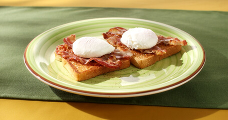 Toast with poached egg and bacon on a plate. Serving breakfast on a colored background. Boiled eggs on bread with fried bacon. Angle view. Sunny day.