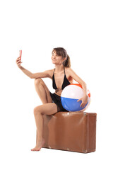 Happy girl in bikini taking selfy photo with colorful inflatable beach ball sitting on big vintage bag. Summer travel concept.