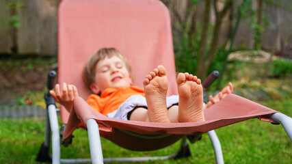 Close up of kid's feet, barefoot , on patio chair. Cute happy little boy relaxing on the chaise...