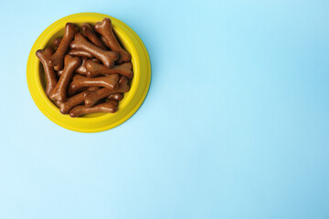 Yellow bowl with bone shaped dog cookies on light blue background, top view. Space for text