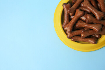 Yellow bowl with bone shaped dog cookies on light blue background, top view. Space for text