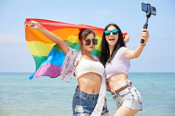 LGBT lesbian couple with a rainbow flag and taking selfie from smartphone on the beach