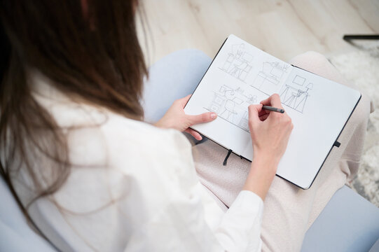 A young woman illustrator, dressed in blue jeans and a white shirt, creates a new illustration by drawing in her sketchbook. She is engrossed in her artwork, bringing her sketches to life.