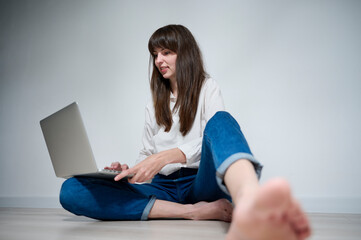 An attractive girl sitting on the floor works remotely on her laptop, responding to various...