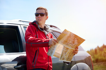 A man leans against a car, holding a map in his hands, as he looks for directions. The traveler...