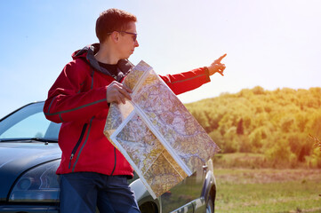 A man leans against a car, holding a map in his hands, as he looks for directions. The traveler...