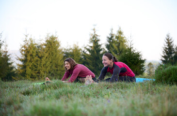 Fototapeta premium Two athletic young women dressed in sporty attire engage in a range of yoga poses amidst the scenic beauty of mountainous surroundings. The gentle rays of the morning sun cast on their slender bodies